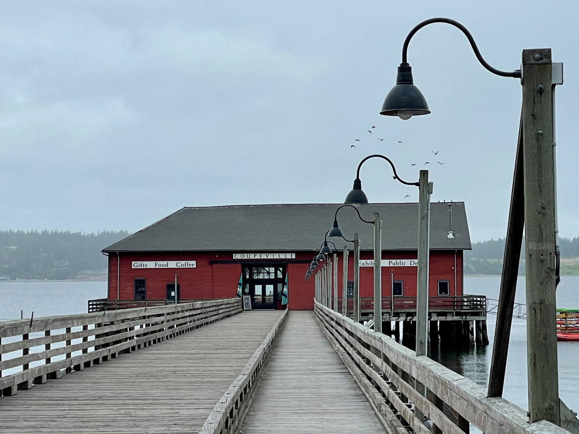 The Coupeville Wharf pictured on an overcast day. The photograh was taken from the front dock of the Wharf. Light posts line the right edge of the dock leading to the Wharf's front door. A flock of birds can be faintly seen above the Wharf.